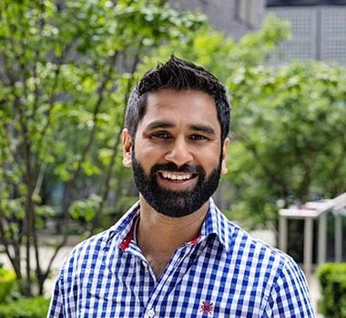 Dr Amit Patel - a man with dark hair and a beard - is smiling at the camera. He's wearing a navy and white gingham shirt. Photo by Charlotte Gray.