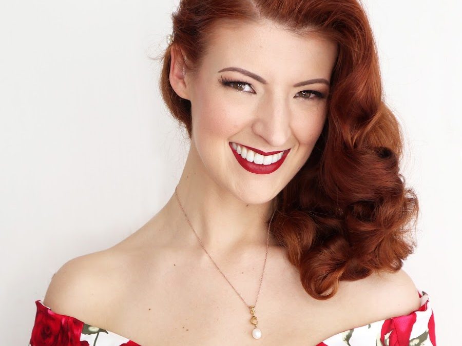 Jessica Kellgren-Fozard - a white woman with auburn hair, wearing a rose-print 1950s style sweetheart neckline dress, with hair styling and lipstick to match.