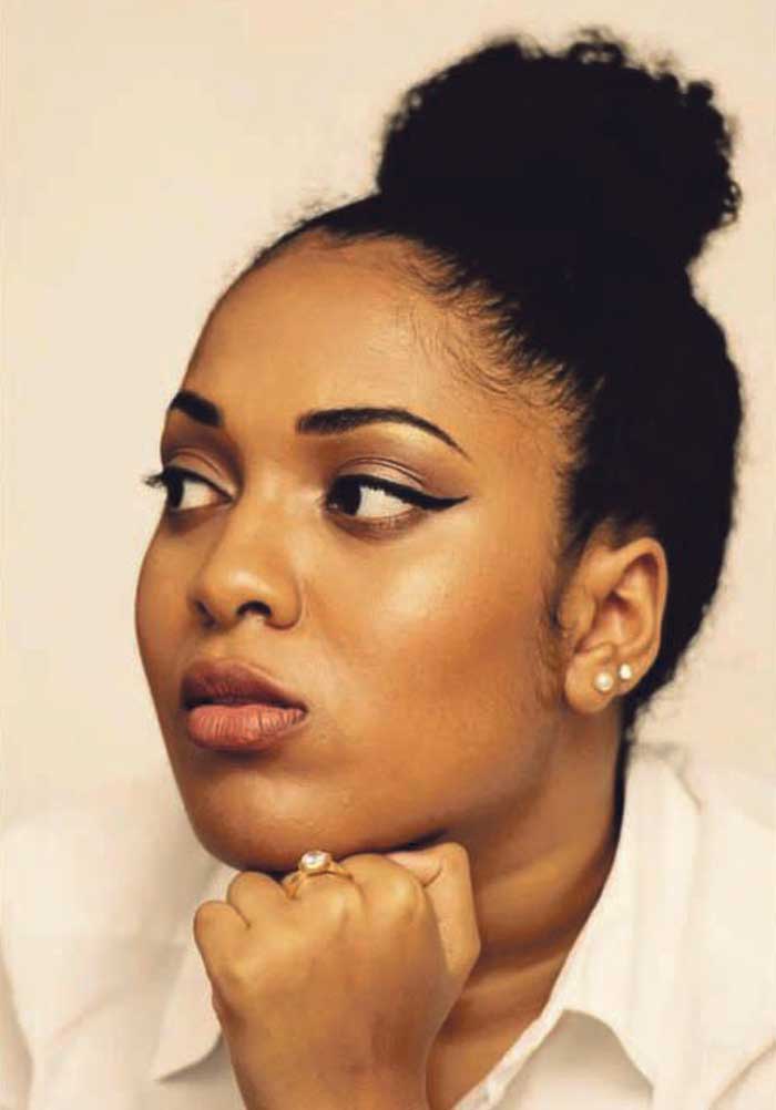 Saida Ahmed - a young Black disabled woman with her hair in a top-knot. She looks off to one side - the photo has a very painterly look to it, with warm tones.