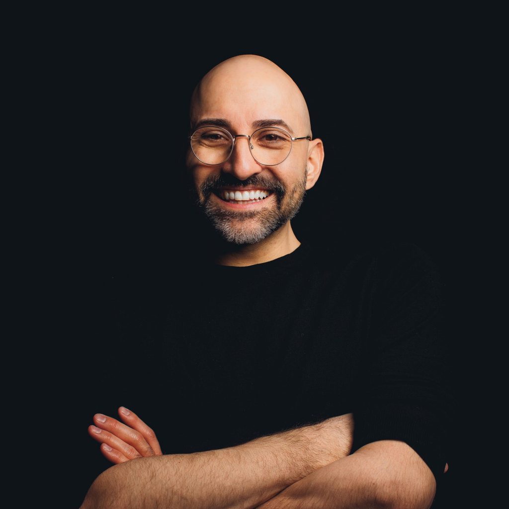 Spencer West -a white man with a broad, sincere smile, a beard and glasses. His arms are folded and he's wearing a black top. Photo by Ryan Bolton