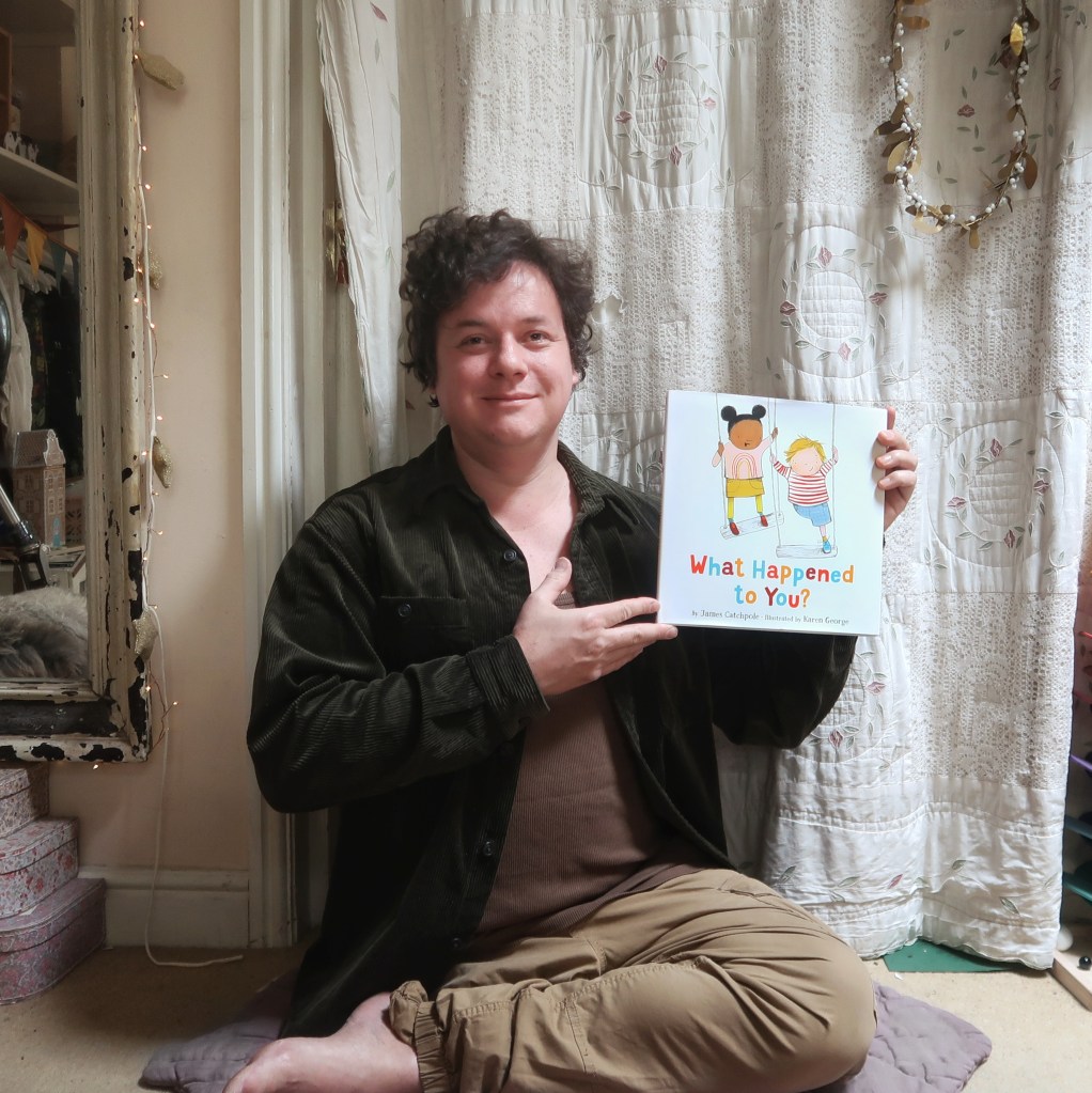 Photo of James Catchpole - a man with one leg and short brown hair - holding the US edition of his picture book, What Happened to You? He's sitting on the floor looking up at the camera.