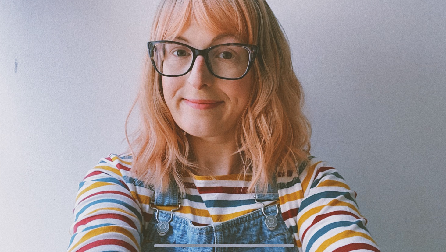 Jen Campbell - a white woman wearing glasses and a pink-tinged wig, looks into the camera with a half-smile. She's wearing denim dungarees and a striped top in off-white, burgundy, mustard and teal.