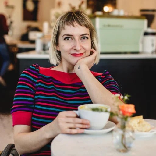 Rebekah Taussig - a white woman with blonde-brown hair is sitting at a cafe table with a hot drink. Her red lipstick matches the red stripe, collar and cuffs of her top, she's smiling warmly at the camera. Photo via A Cup of Jo.