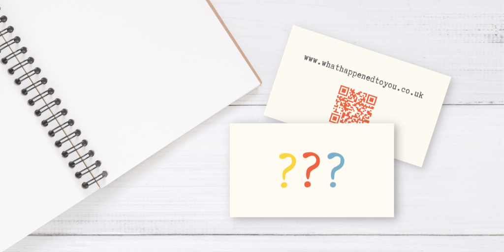 A cream card with 3 question marks. Next to it is the same card with the other side showing, with a QR code and the url www.whathappenedtoyou.co.uk - on a white wood background, lying next to a notebook